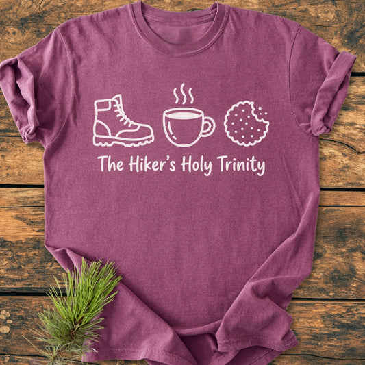 The Hiker's Holy Trinity - Vintage Wash Tee features icons of a hiking boot, mug, and cookie with the phrase below, laid on rustic wood with a pine sprig accent.