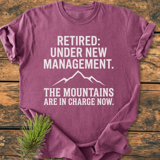 New Management - Vintage Wash Tee in heather-purple, featuring a white graphic with retirement-themed text and mountain silhouette, laid flat on rustic wood, with a pine sprig accent.