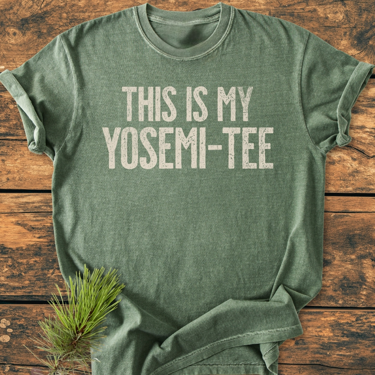 This Is My Yosemi-Tee - Vintage Wash Tee features a forest-green shirt with off-white block letters, laid on wood, evoking an outdoorsy vibe with an accompanying pine branch.
