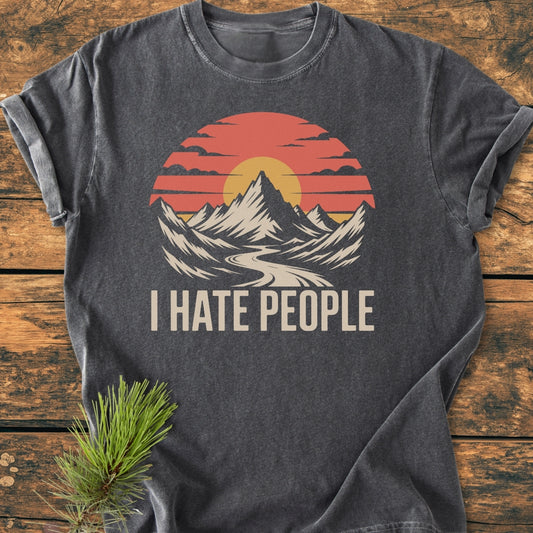 I Hate People - Vintage Wash Tee featuring a retro mountain sunset graphic and bold text, laid on wood with a pine sprig. Short-sleeve, dark-gray, 100% cotton, relaxed fit.
