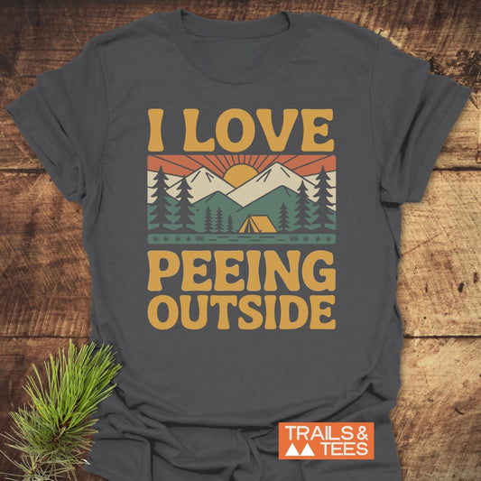 I Love Peeing Outside T-Shirt displays a retro outdoor scene with trees, tent, mountains, and sun, plus “TRAILS & TEES” logo, designed for comfort and adventure in soft ring-spun cotton.