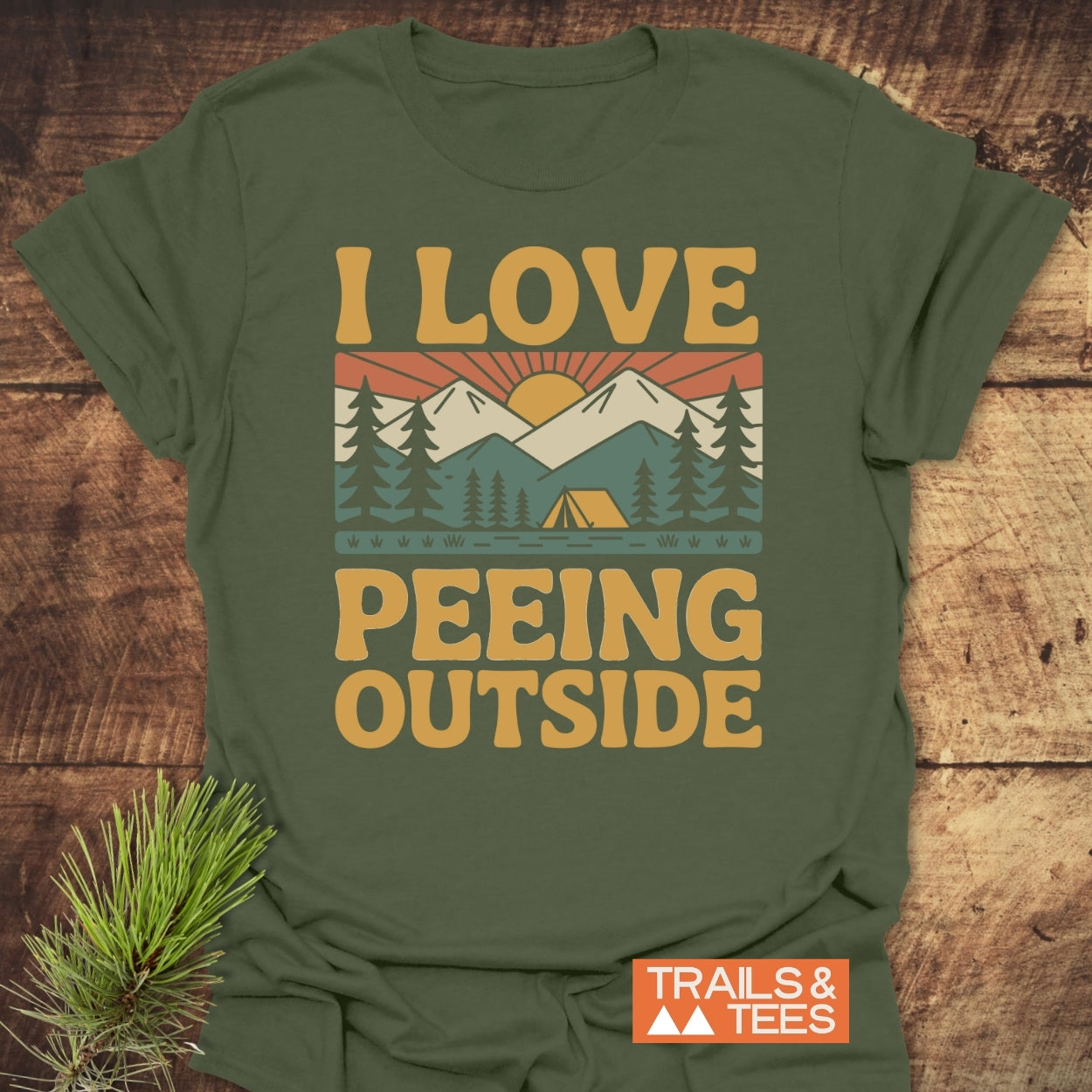 I Love Peeing Outside T-Shirt featuring a stylized outdoor scene with mountains and a tent, crafted from soft ring-spun cotton. Includes 'TRAILS & TEES' logo, laid on a wooden surface.