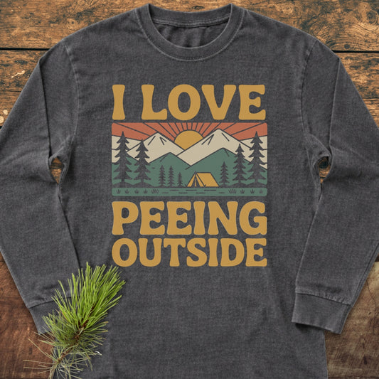 I Love Peeing Outside Long-Sleeve Tee featuring a retro sunrise graphic with mountains and pine trees, laid flat on wood, showcasing its soft, 100% ring-spun cotton material and relaxed fit.