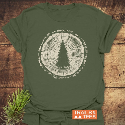 Rooted In Nature T-Shirt featuring a tree trunk cross-section and pine silhouette, laid on wood with a pine sprig. TRAILS & TEES branding visible, emphasizing the outdoors theme.