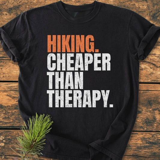 Hiking Cheaper Than Therapy - Vintage Wash Tee: Black t-shirt with orange and white slogan, laid on wood, featuring a small pine sprig. Made from soft, durable, 100% ring-spun cotton for comfort.