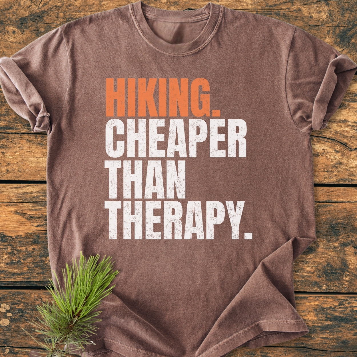 Hiking Cheaper Than Therapy - Vintage Wash Tee displayed on a wooden surface, featuring bold orange and white slogan text. A pine sprig lies beside the soft, durable, 100% ring-spun cotton shirt.