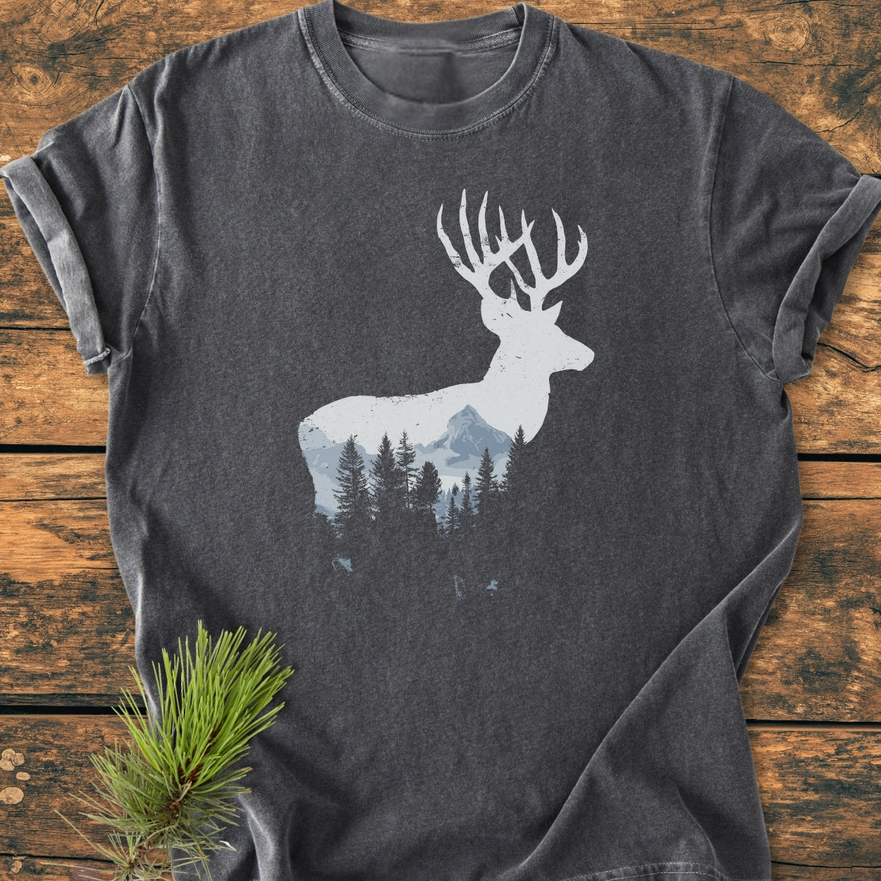 Deer Silhouette - Vintage Wash Tee features a white stag silhouette with a mountain and pine tree scene on a charcoal-gray T-shirt, displayed on a wooden surface with a pine branch accent.