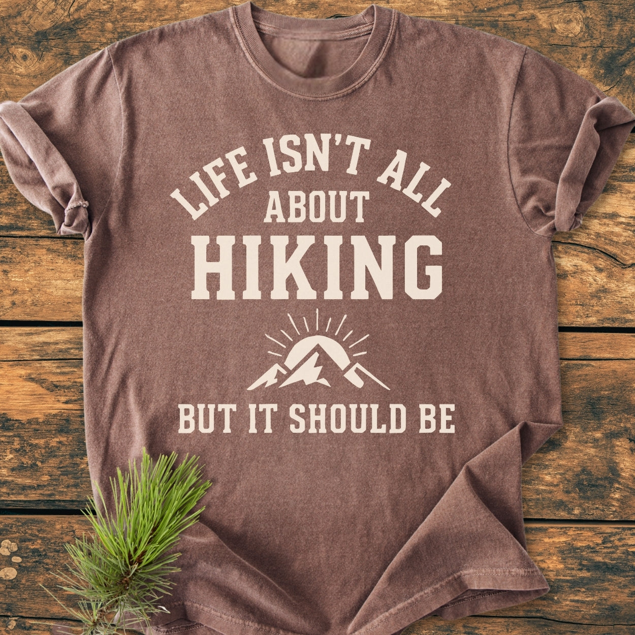 Life Isn't All About Hiking - Vintage Wash Tee with a mountain and sun graphic, laid on wood. Features off-white slogan, pine sprig accent, showcasing its relaxed fit and soft-washed texture.