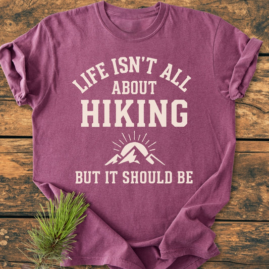 Life Isn't All About Hiking - Vintage Wash Tee, heathered purple, laid flat on wood with pine sprigs. Features bold white slogan and mountain graphic, highlighting its relaxed fit and durable construction.