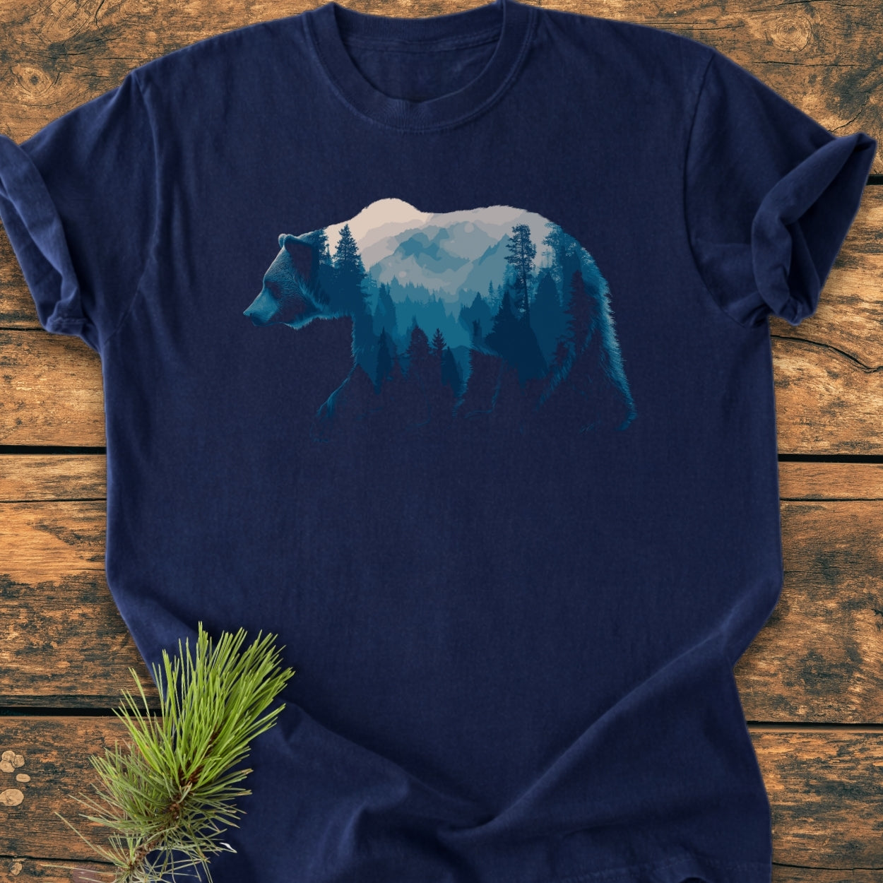 Grizzly Silhouette - Vintage Wash Tee, featuring a stylized bear with a layered forest and mountain scene, laid on wood with a pine sprig. Crafted from soft, durable, 100% ring-spun cotton.