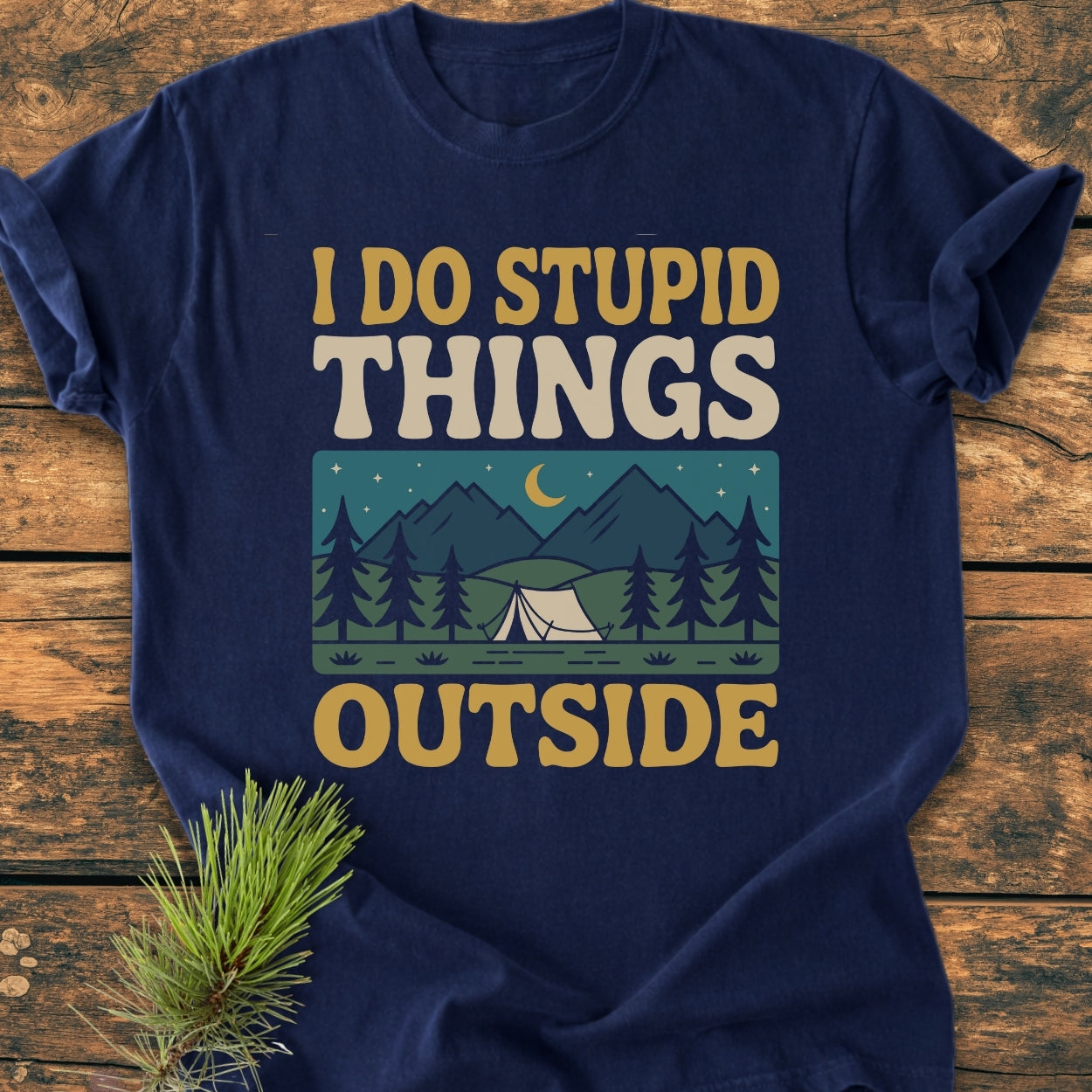 I Do Stupid Things Outside - Vintage Wash Tee features retro lettering and a night camping scene illustration on a navy-blue T-shirt, laid on a wooden surface with a pine sprig beside it.