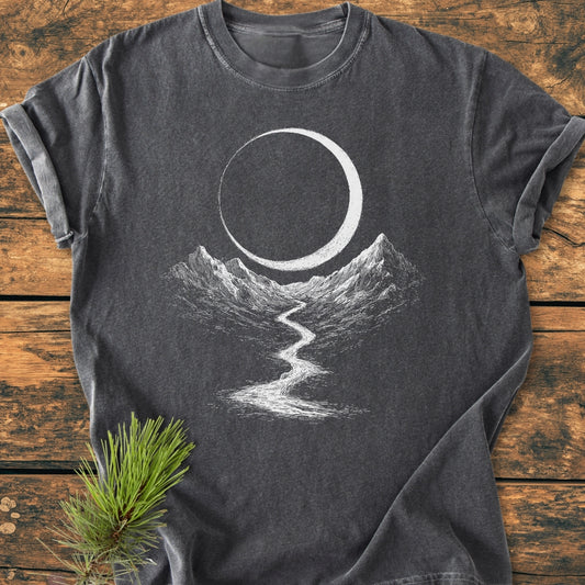 Eclipse Trail - Vintage Wash Tee: Charcoal-gray T-shirt with white line art of mountains, river, and crescent moon, laid flat on wood with pine sprig. Made of soft, 100% ring-spun cotton.