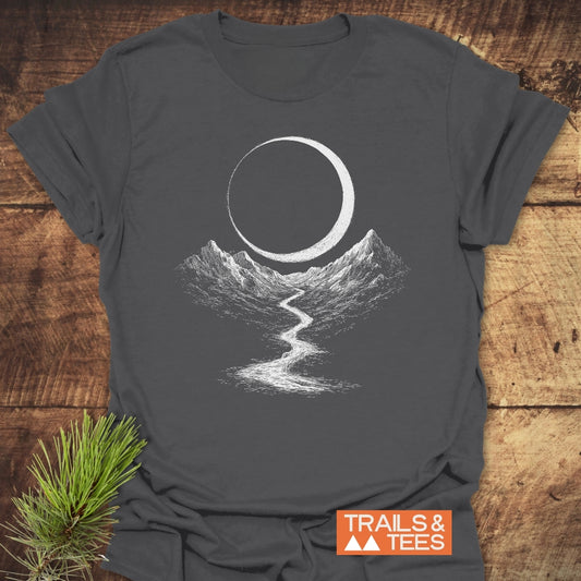 Eclipse Trail T-Shirt featuring a white graphic of mountains and a trail, symbolizing outdoor adventure, on a charcoal-gray fabric. Laid on a wooden surface, with the TRAILS & TEES logo displayed.