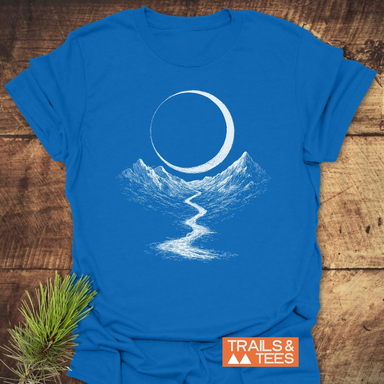 Eclipse Trail T-Shirt: Bright blue tee featuring a white nature scene with mountains, river, and crescent moon. Laid on wood, it embodies outdoor adventure. Includes TRAILS & TEES brand logo.