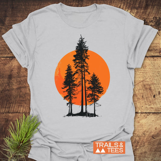 Sunset Trees T-Shirt featuring black pine-tree silhouettes against an orange circle, laid on wood. Made from soft ring-spun cotton, it embodies trail-tested comfort and adventure-ready design, showcasing the TRAILS & TEES logo.