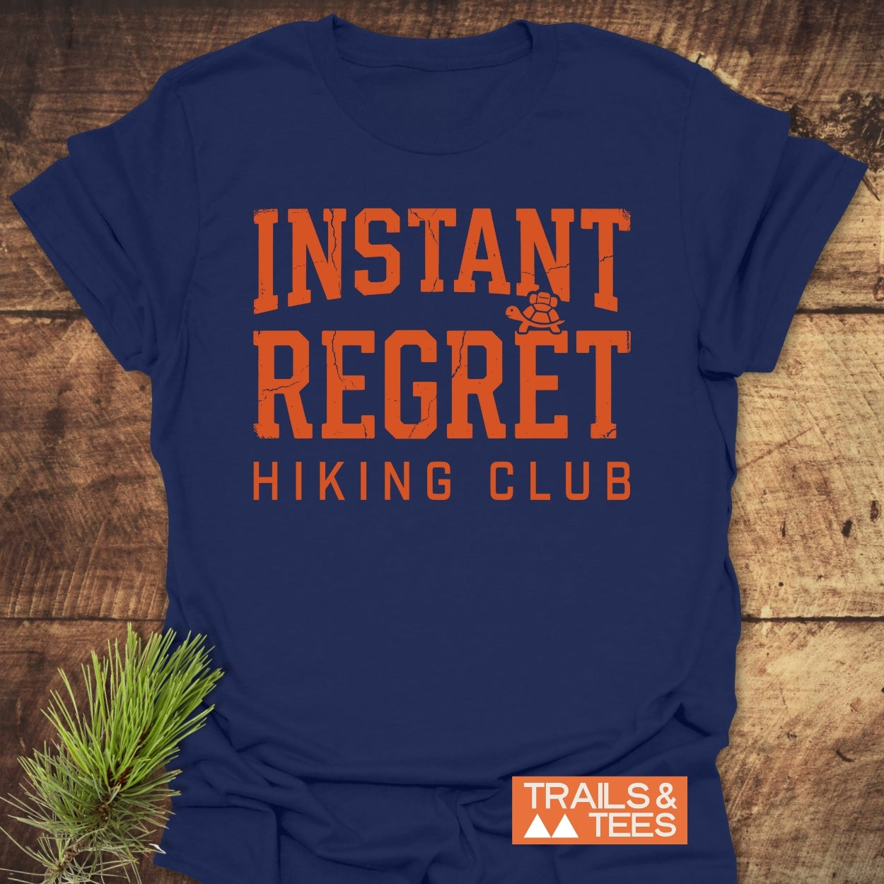 Navy-blue Instant Regret Hiking Club T-shirt with orange lettering and a turtle icon, featuring the TRAILS & TEES logo. Laid on wood with a pine sprig beside it.