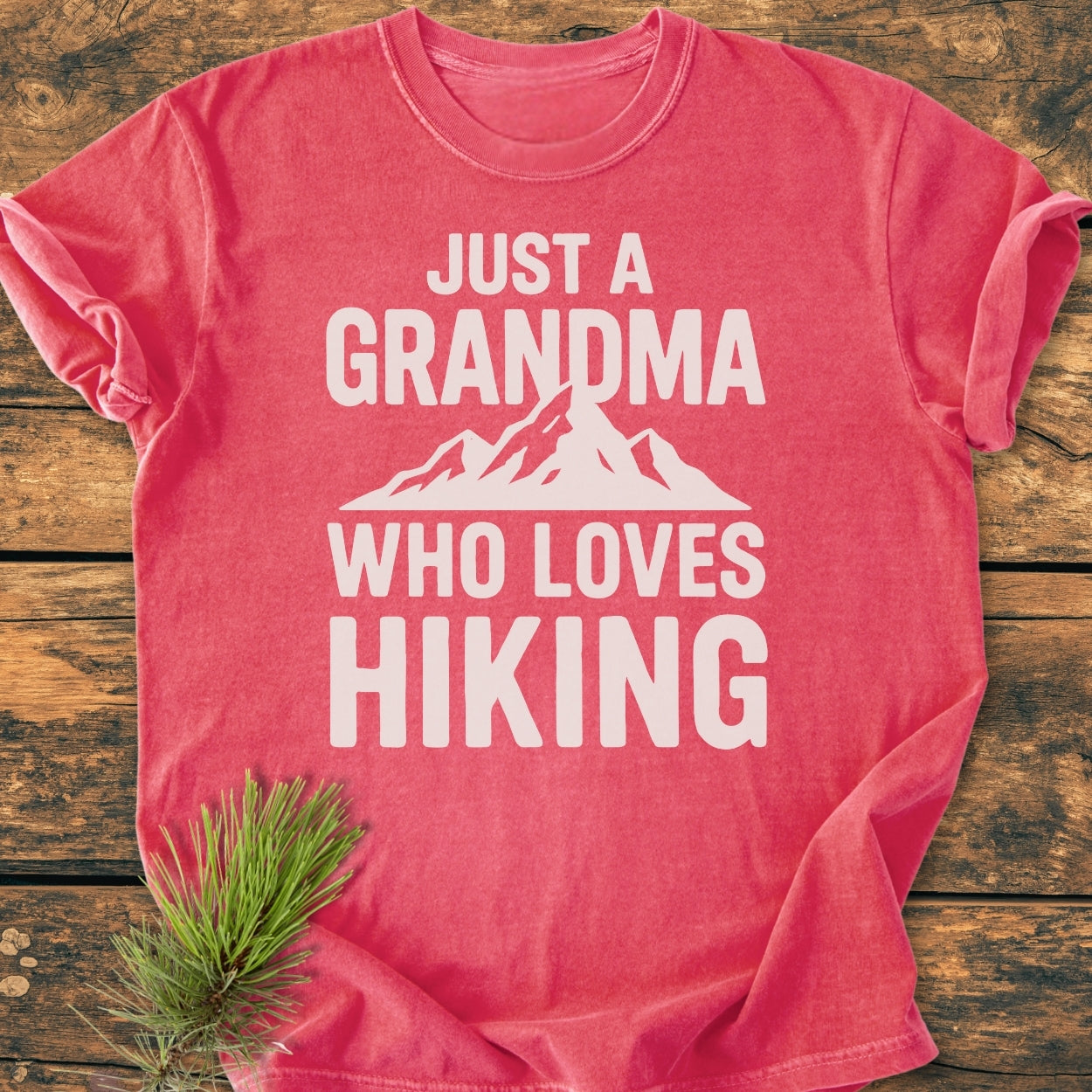 Coral-pink Grandma Loves Hiking - Vintage Wash Tee with white text and mountain graphic, displayed flat on wood surface, accompanied by an evergreen branch, highlighting its casual, relaxed fit and cozy fabric.