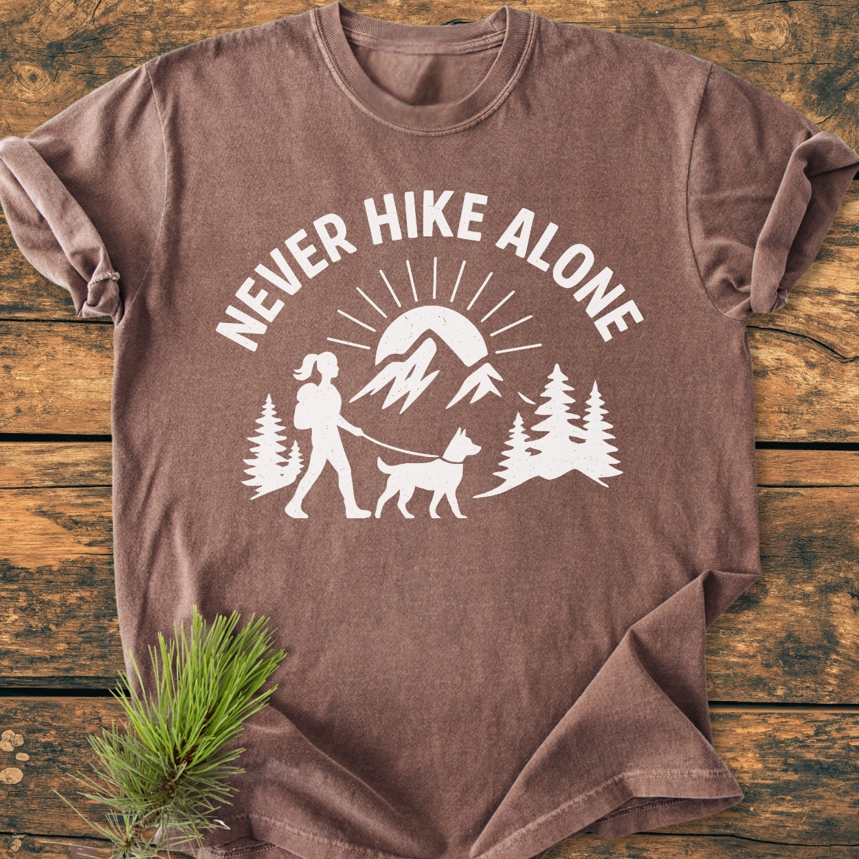 Never Hike Alone Dog Mom - Vintage Wash Tee features a white graphic of a hiker with a dog and mountains, emphasizing adventure. Laid on wood, it highlights the relaxed, cozy fit.