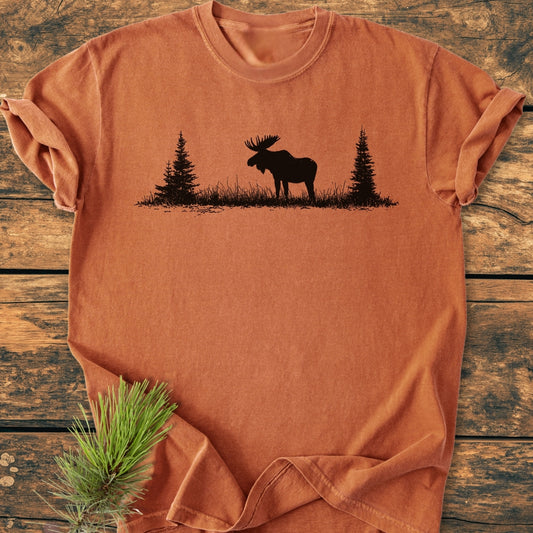 Lone Moose - Vintage Wash Tee features a black moose and pine tree silhouette on a rust-orange, short-sleeve T-shirt, laid on a wooden surface. Cozy and durable with a relaxed fit.