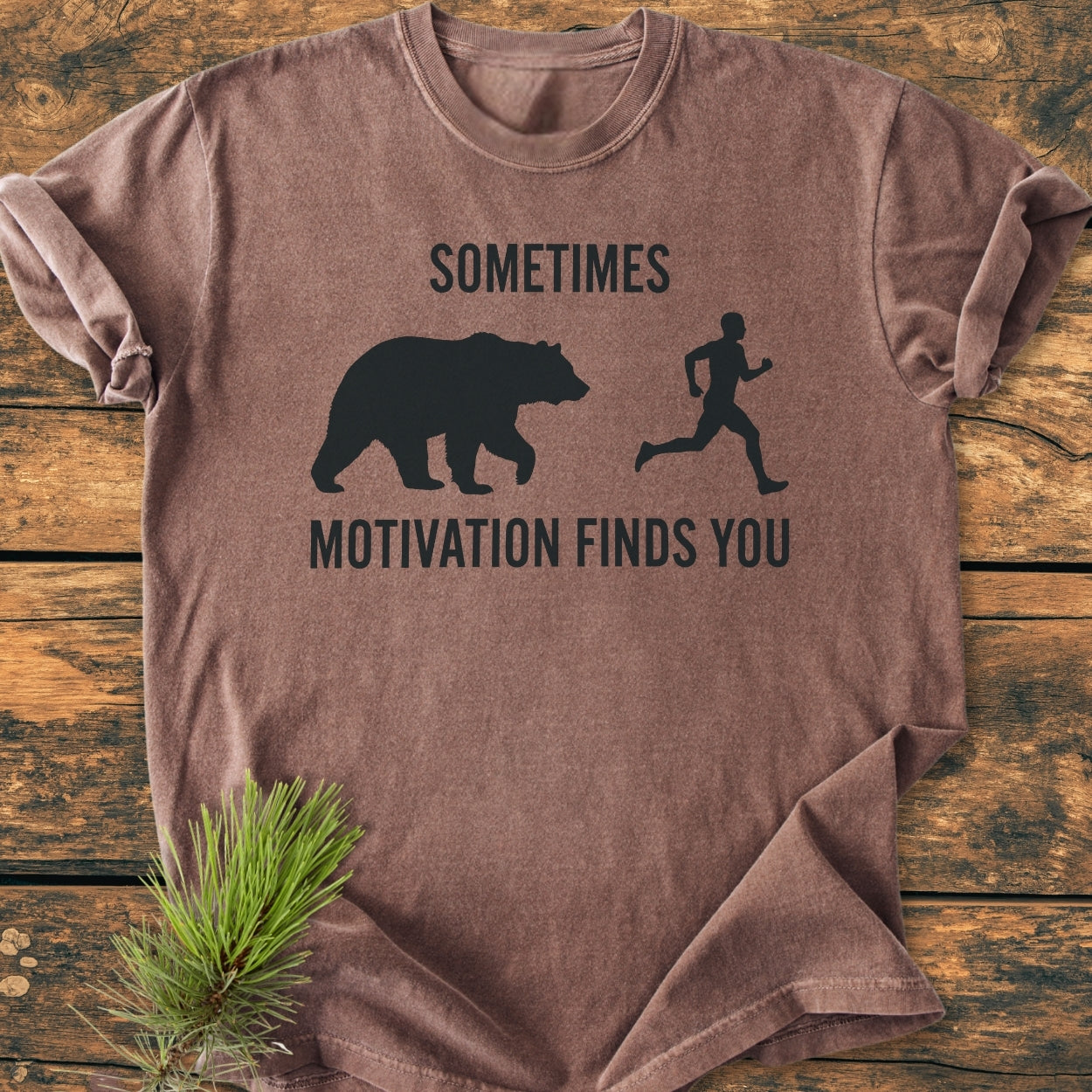 Sometimes Motivation Finds You - Vintage Wash Tee displayed on a rustic wooden surface, featuring a black bear and runner graphic with the slogan. Made of 100% ring-spun cotton for comfort and durability.