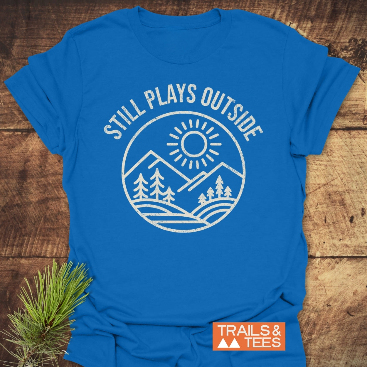 Still Plays Outside T-Shirt featuring a white sun and mountain graphic, laid on wood. Includes “TRAILS & TEES” logo, embodying outdoor adventure with comfort and durability.