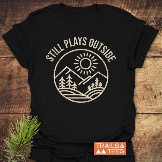 Still Plays Outside T-Shirt featuring a white sun and mountain design with pine trees on black fabric. Includes an orange TRAILS & TEES label, ideal for outdoor enthusiasts.