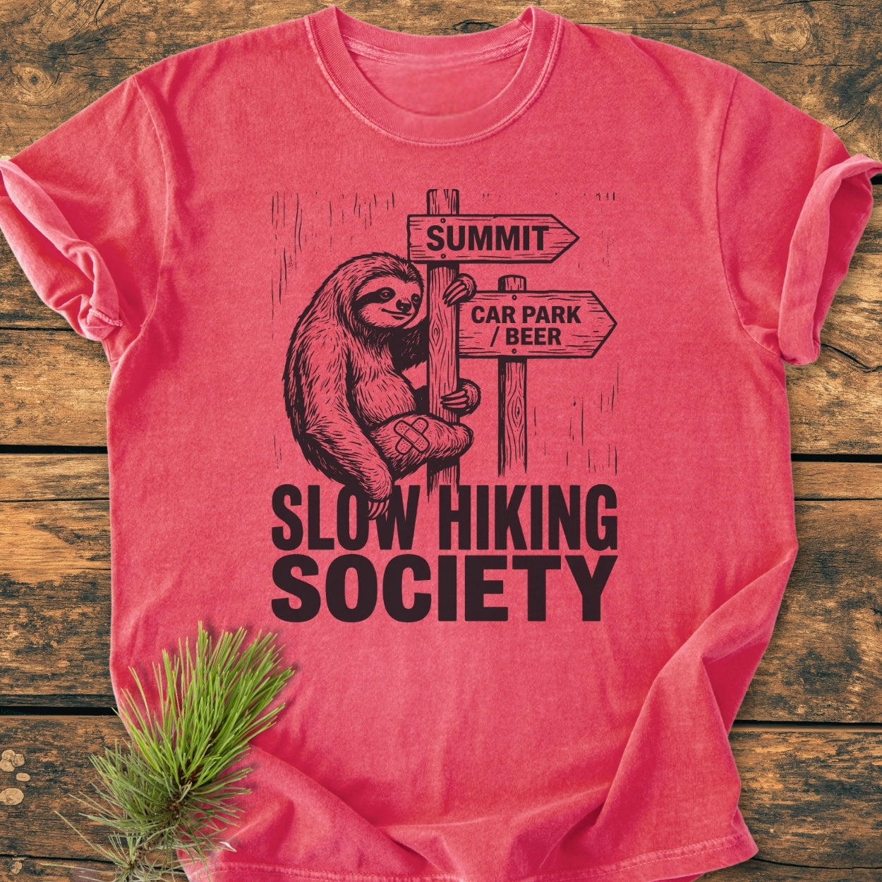 Flat-lay of the Slow Hiking Society - Vintage Wash Tee, featuring a cartoon sloth and humorous signpost design, laid on a wooden surface with a pine sprig, highlighting its casual, relaxed style.