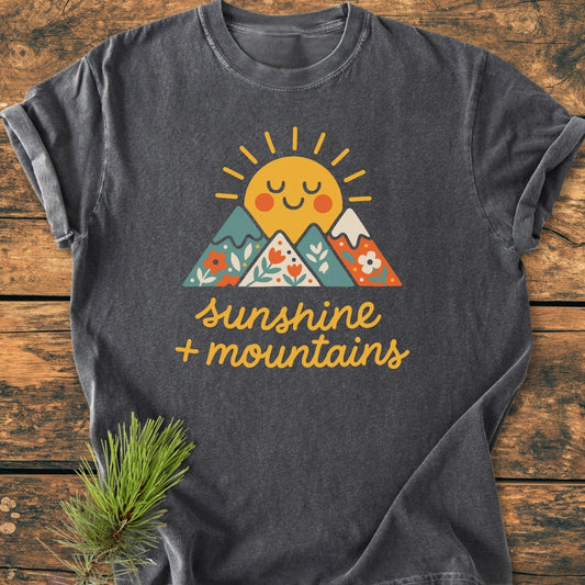 Sunshine And Mountains - Vintage Wash Tee features a cheerful sun graphic behind flower-patterned mountains with sunshine + mountains text, on a dark-grey, short-sleeve tee, laid on a wooden surface.