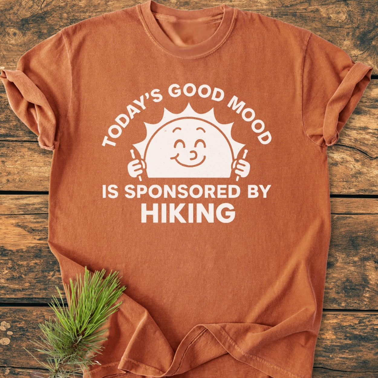 Today's Good Mood - Vintage Wash Tee features a rust-orange color with a smiling sun and hiking slogan, styled with a pine sprig on a wood surface.