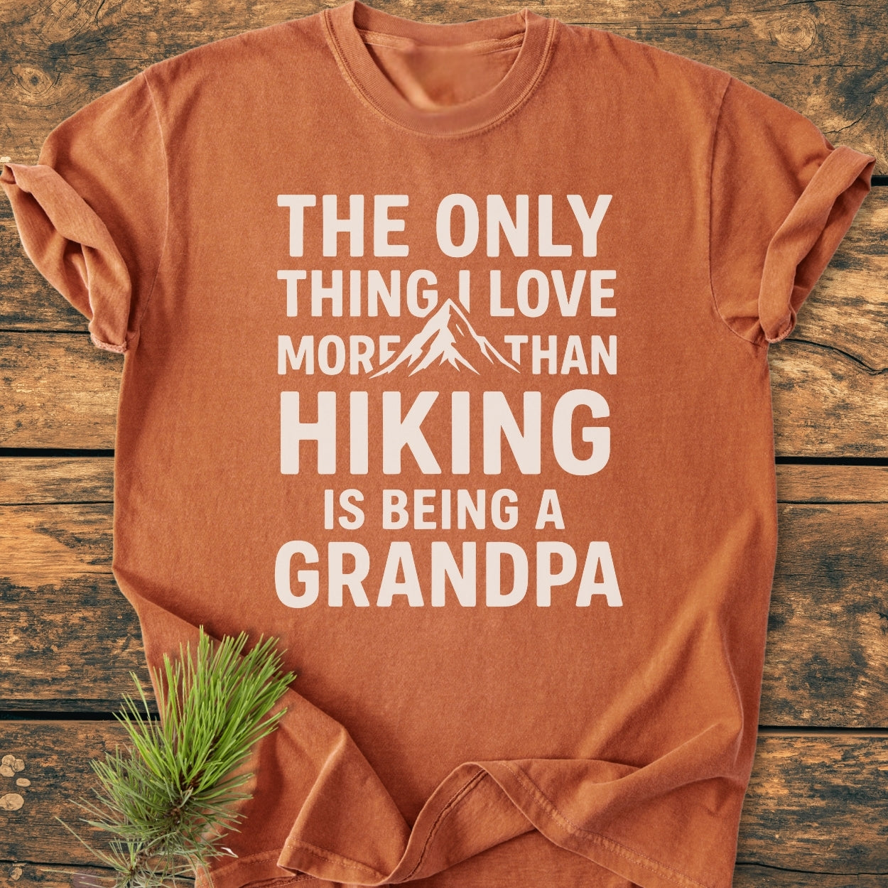 Rust-orange Hiking Grandpa - Vintage Wash Tee with white block letters and mountain graphic, emphasizing hiking and grandpa love, laid flat on wood. Pine needles accent the scene.