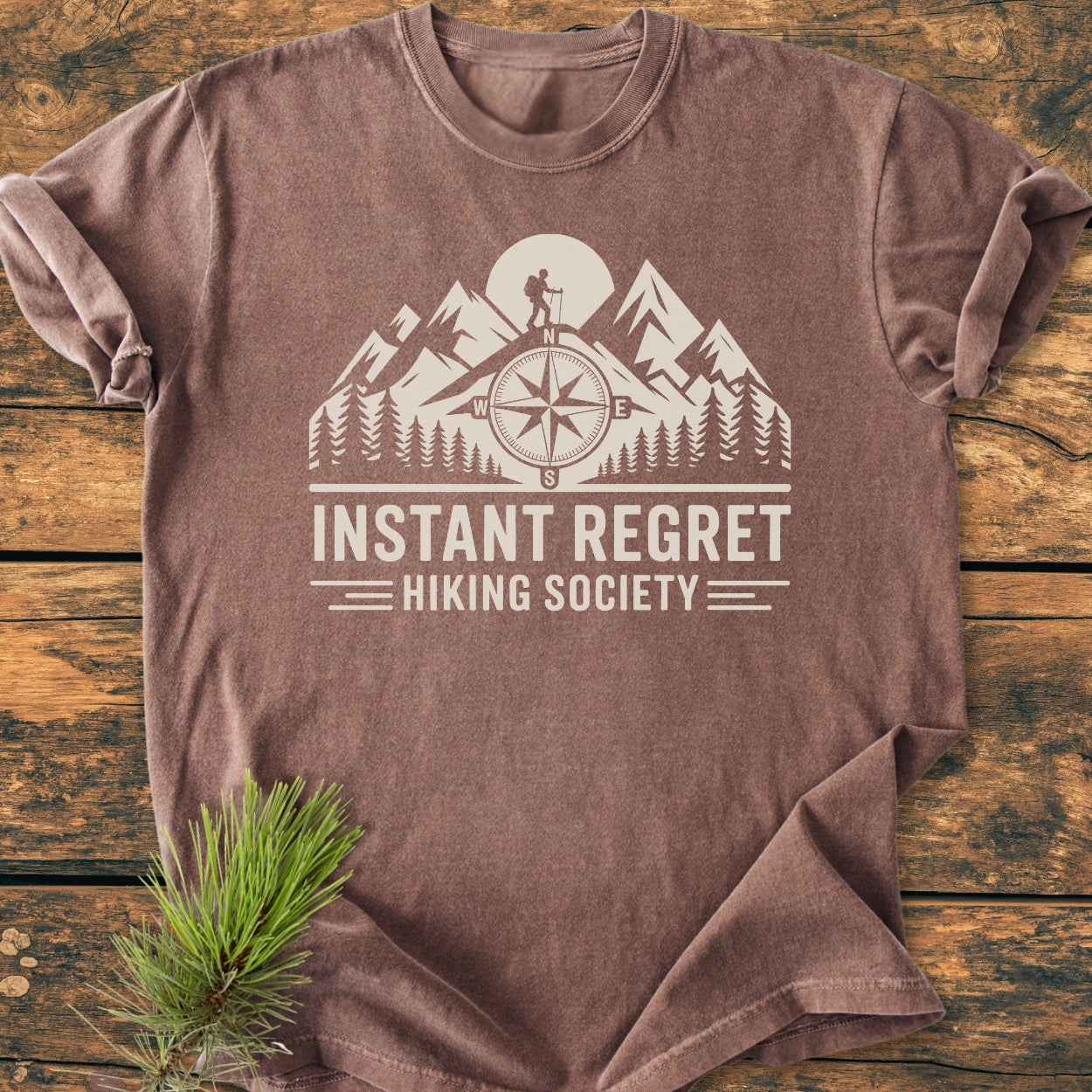 Instant Regret Hiking Society - Vintage Wash Tee features a white graphic of mountains, a compass, and a hiker on a brown, short-sleeve T-shirt, styled with a pine sprig.