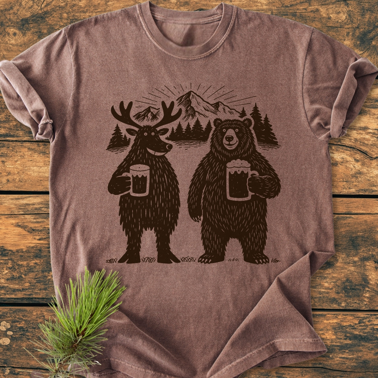 Bear, Deer And Beer - Vintage Wash Tee features a cartoon moose and bear with beer mugs, set against pine trees and mountains, laid on a wooden surface with pine needles for styling.