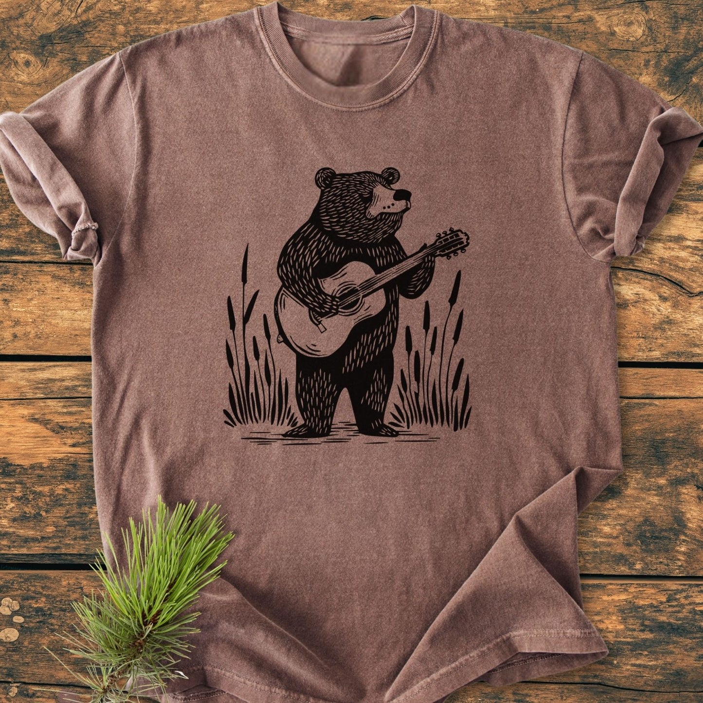 Bear Playing Guitar - Vintage Wash Tee featuring a bear with a guitar amid reeds on a brown, garment-dyed cotton. Laid flat on wood, showcasing its relaxed fit and rolled-up sleeve.