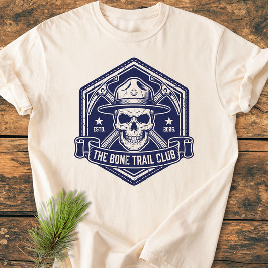 The Bone Trail Club - Vintage Wash Tee features a hexagonal skull badge with a ranger hat, crossed ice-axes, and THE BONE TRAIL CLUB banner. Laid on rustic wood with pine sprig.