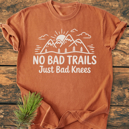 No Bad Trails - Vintage Wash Tee featuring a mountain design with a sun, trees, and slogan on a rust-orange short-sleeve laid on wood, accompanied by a pine sprig.
