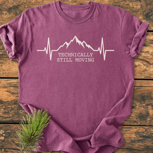 Still Moving - Vintage Wash Tee in heathered purple, featuring a stylized heartbeat line transitioning into mountains with “TECHNICALLY STILL MOVING” text. Displayed on rustic wood with a pine sprig accent.