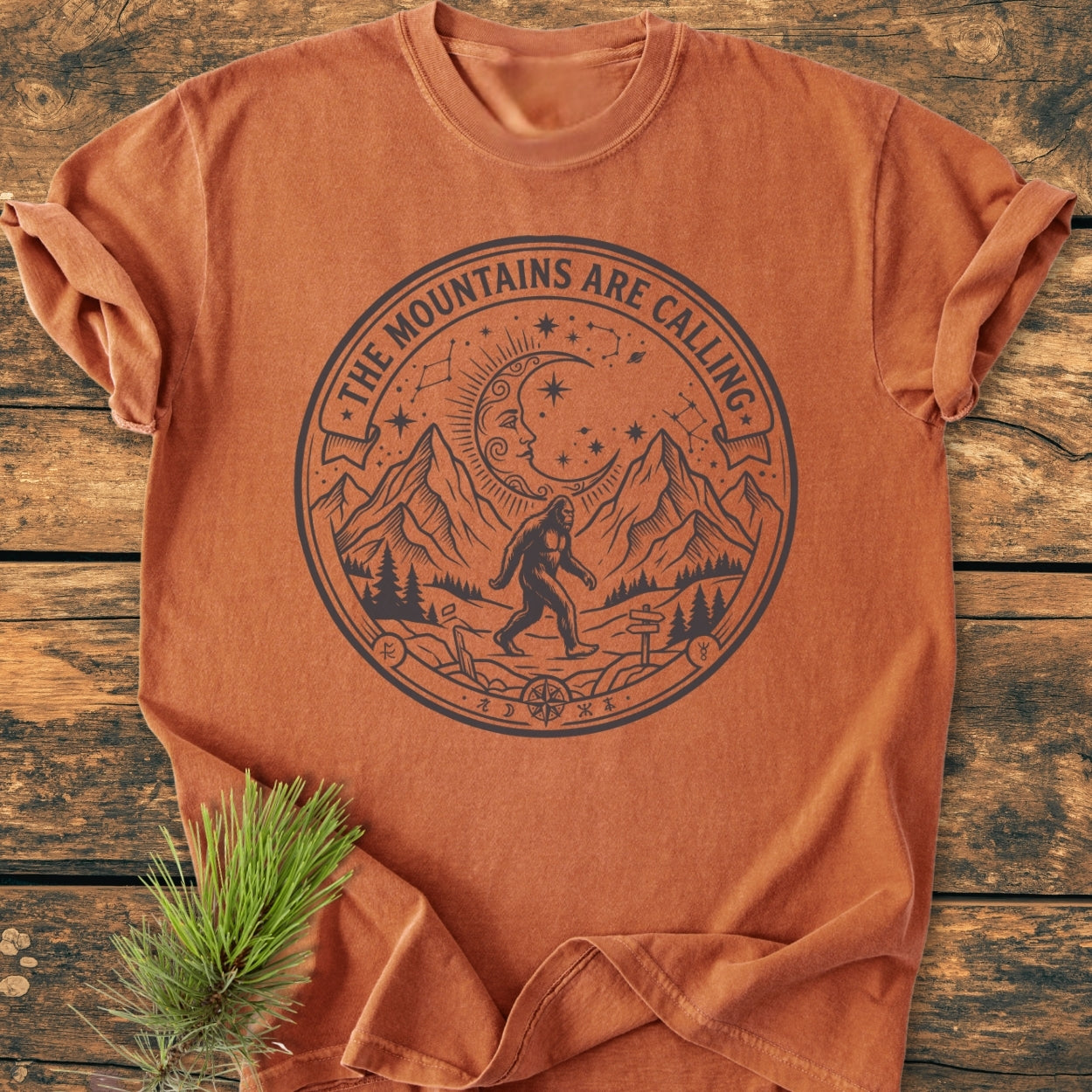 The Mountains Are Calling - Vintage Wash Tee features a wilderness scene with Bigfoot on a burnt-orange T-shirt, laid on wood, accented by a pine sprig for rustic flair.