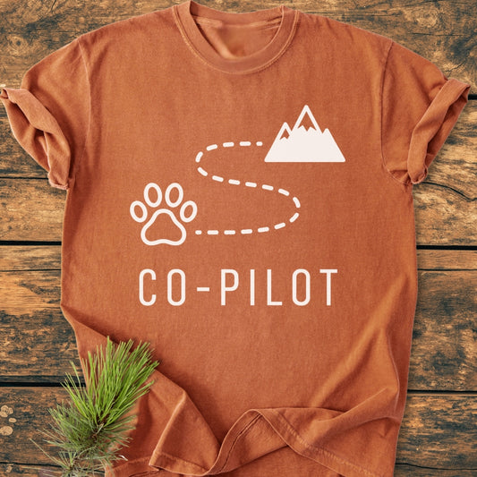 Co-Pilot Dog - Vintage Wash Tee: Rust-orange short-sleeve T-shirt featuring a white paw print and mountain graphic with CO-PILOT text, laid flat with a pine sprig for an outdoorsy touch.