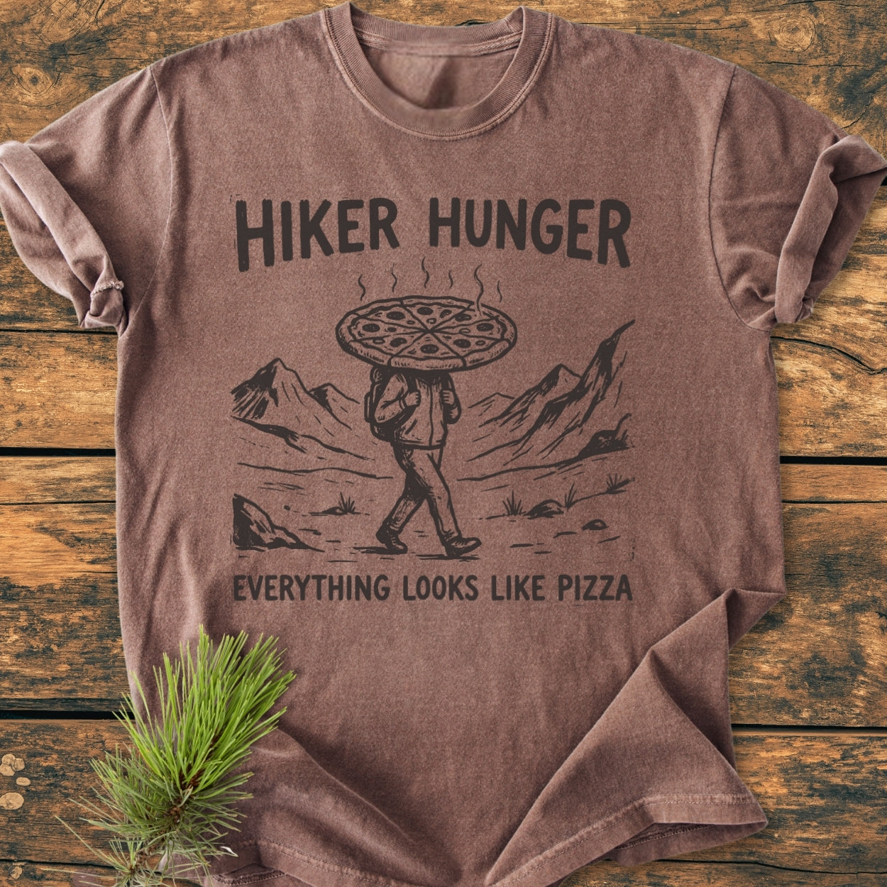 Hiker Hunger - Vintage Wash Tee featuring a stylized hiker with a pizza head and mountain peaks graphic. Laid on wood with a pine sprig, showcasing soft, durable garment-dyed cotton.