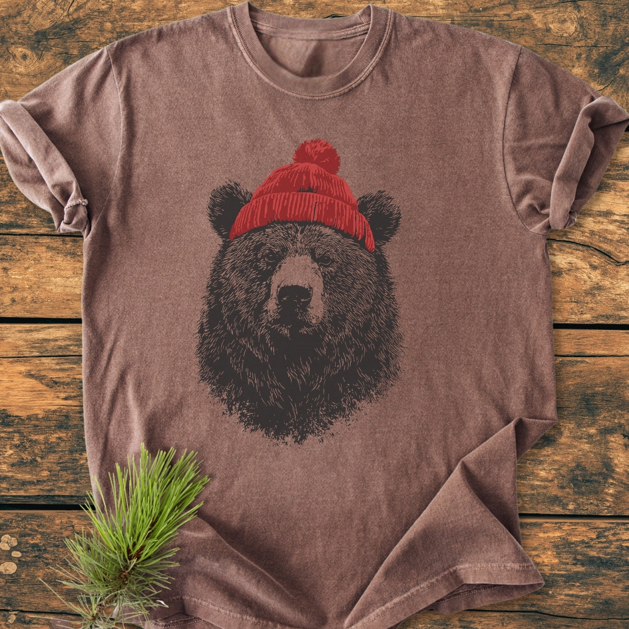 Bear With Beanie - Vintage Wash Tee: A muted brown, short-sleeve T-shirt featuring a bear graphic wearing a red knit beanie, laid flat on wood with pine needles beside it.