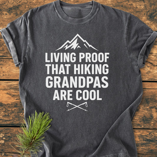 Living Proof Grandpa - Vintage Wash Tee features a bold mountain design with hiking poles on a charcoal-gray T-shirt, laid on wood, complemented by a pine sprig for an outdoorsy touch.