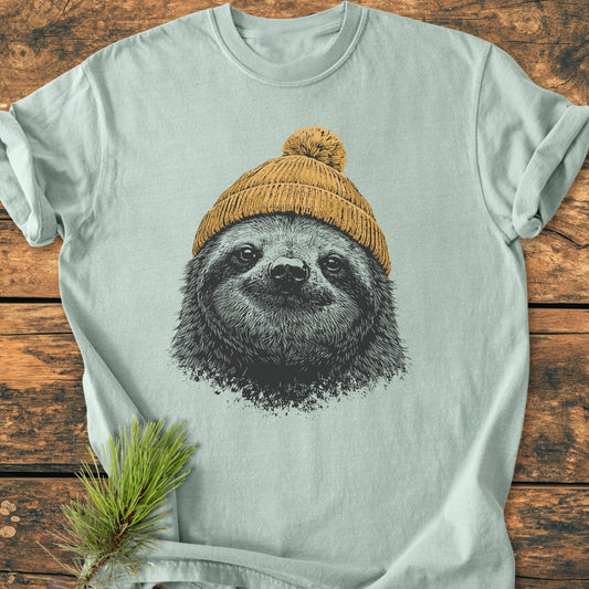 Sloth With Beanie - Vintage Wash Tee features a pastel green design with a sloth in a mustard-yellow beanie, laid on rustic wood, highlighting its relaxed fit and soft-washed texture.