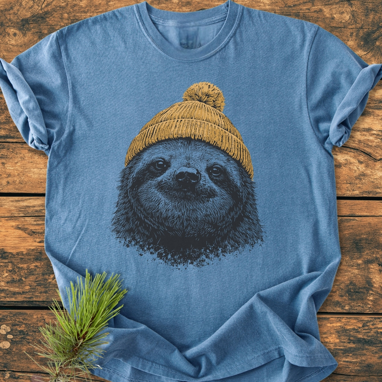 Sloth With Beanie - Vintage Wash Tee features a stylized sloth face in dark ink wearing a mustard-yellow beanie. Laid flat on wood, showcasing the soft, relaxed fit of the garment-dyed fabric.