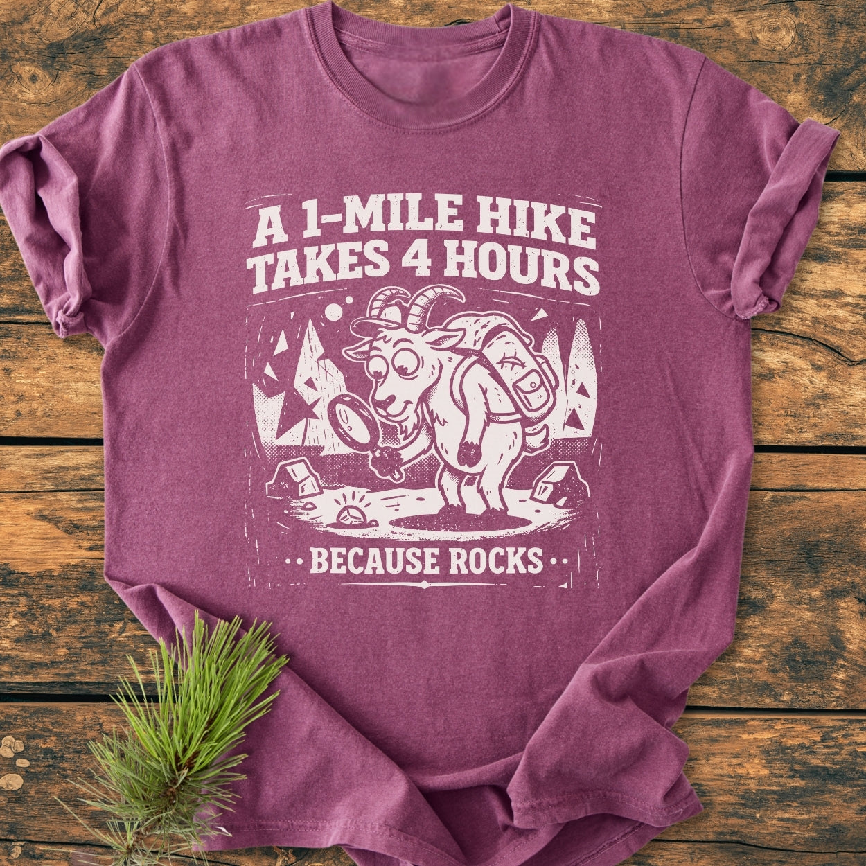 Because Rocks - Vintage Wash Tee features a cartoon hiking goat with a magnifying glass on a purple-mauve T-shirt, highlighting the slogan: “A 1-Mile Hike Takes 4 Hours … Because Rocks.”