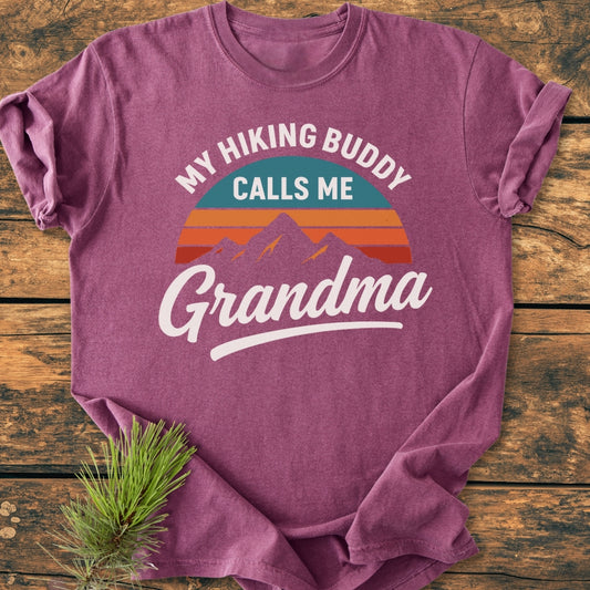 Hiking Buddy Grandma - Vintage Wash Tee: A mauve T-shirt featuring a retro sunset and mountain graphic with the slogan. Laid flat on wood, adorned with a pine sprig accent.