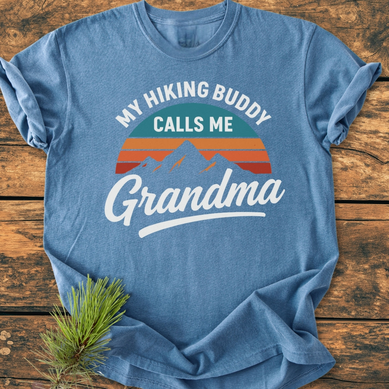 Hiking Buddy Grandma - Vintage Wash Tee in heather-blue, featuring a retro mountain graphic and playful text. Laid on wood with pine sprig, showcasing the relaxed fit and durable stitching.