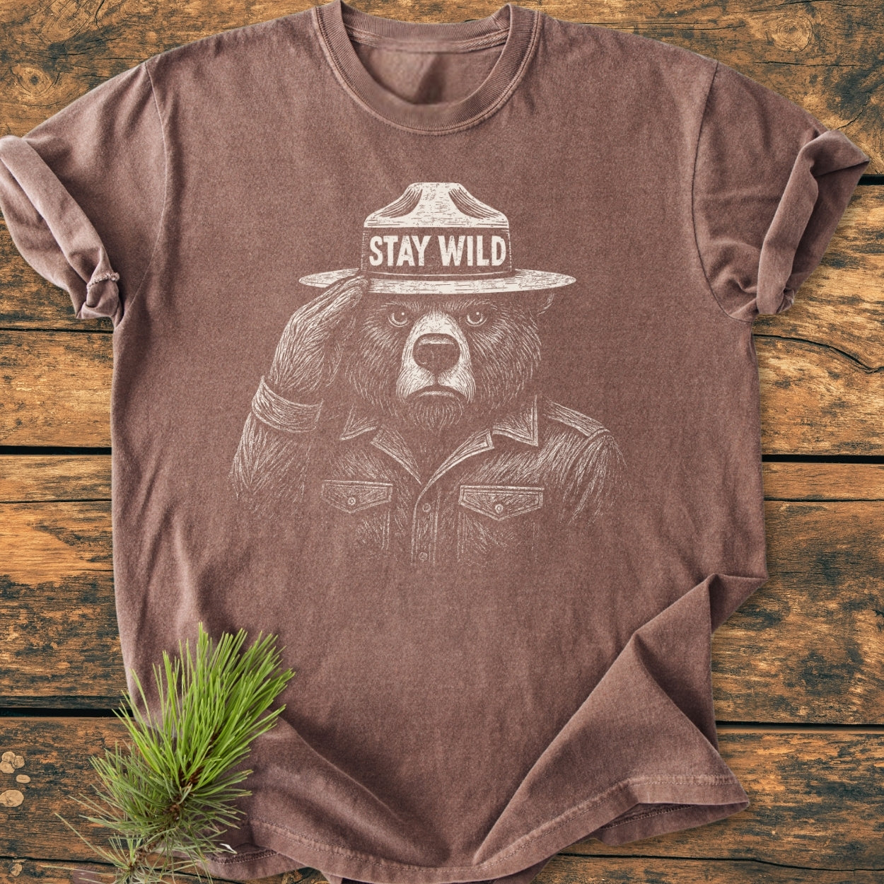 Stay Wild - Vintage Wash Tee features a brown short-sleeve design with a hand-drawn bear in a ranger hat saluting on the front, laid on a wooden surface with a pine sprig nearby.