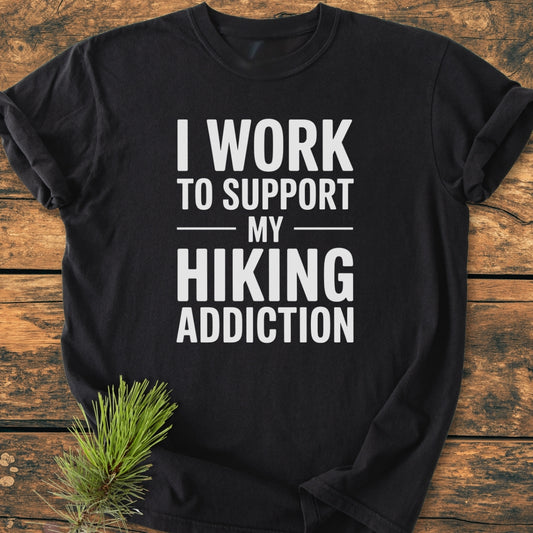 Hiking Addiction - Vintage Wash Tee, a black short-sleeved shirt with a bold hiking slogan, lies on rustic wood, adorned with a pine sprig, showcasing its outdoor spirit and cozy, relaxed fit.