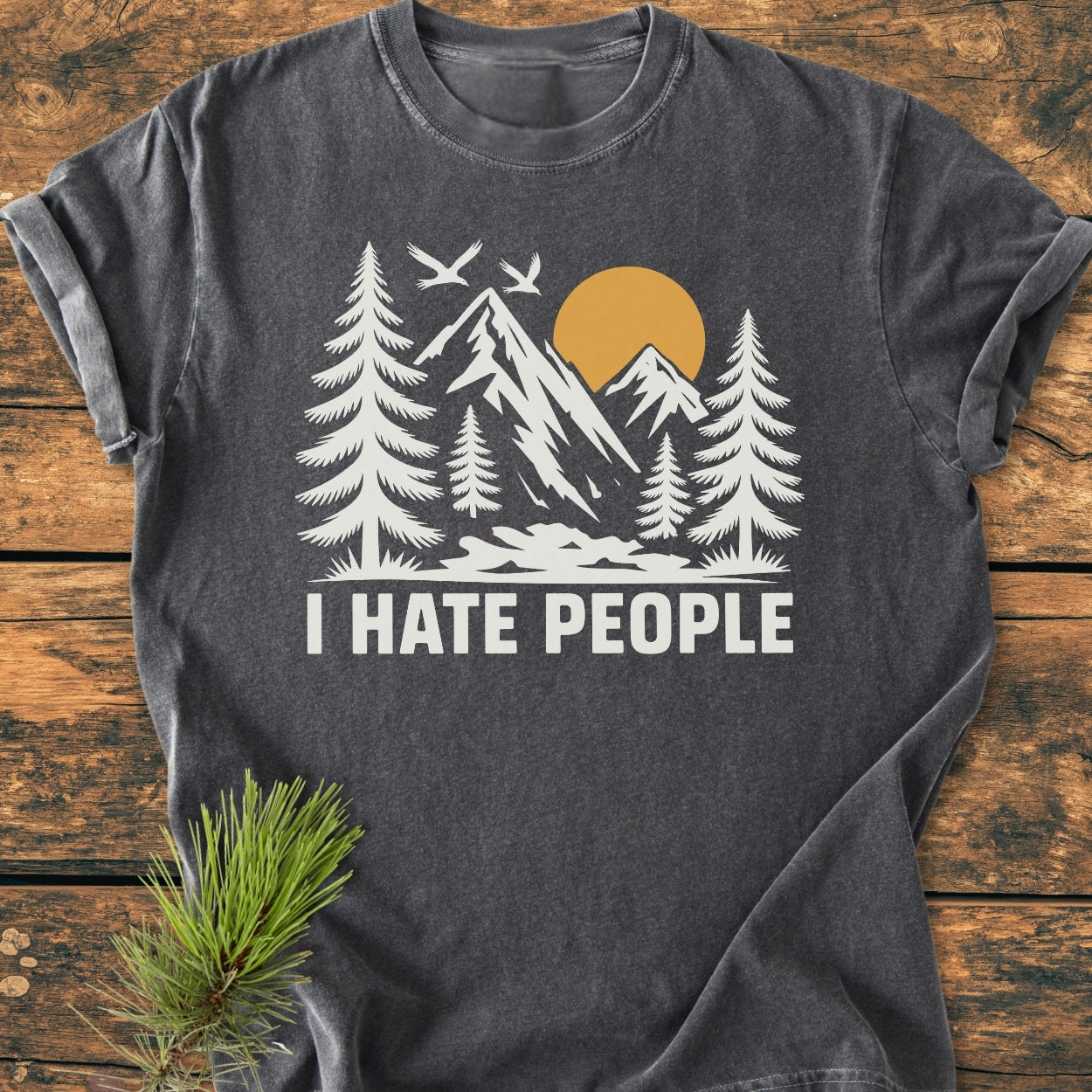 I Hate People - Vintage Wash Tee features a dark gray base with a white and muted gold nature scene, highlighting pine trees and mountains. Laid flat with a pine sprig accent.