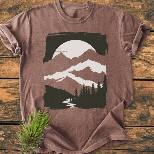 Minimalist Mountains - Vintage Wash Tee featuring a nature scene with mountains, sun, pine trees, and a river, laid flat on wood. Includes pine sprig prop, highlighting its cozy, relaxed design.